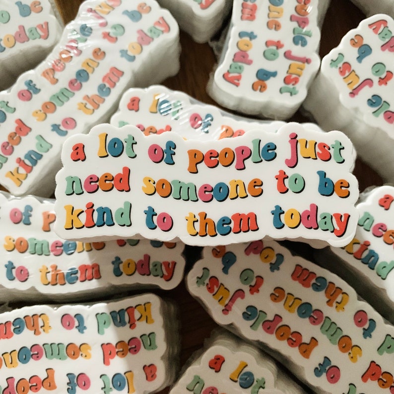 Op de afbeelding: Een stapel kleurrijke stickers met de tekst "a lot of people just need someone to be kind to them today" erop gedrukt. De stickers zijn wit met regenboogkleurige letters.