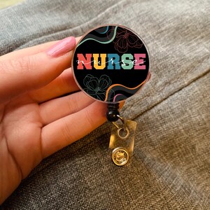 Könnte beinhalten: Ein schwarzer und weißer ausziehbarer Badgehalter mit einem bunten "NURSE"-Design. Das Design zeigt weiße Blumen und wellenförmige Linien auf schwarzem Hintergrund.