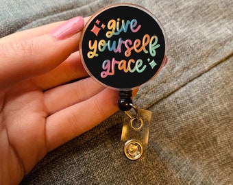 May include: A black and white round badge holder with a rainbow colored text that reads "give yourself grace" and two diamond shapes on either side of the text. The badge holder is attached to a black and silver retractable badge reel.