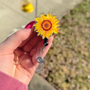 以下が含まれることがあります： 茶色の芯を持つ黄色のひまわりが透明なプラスチック製のバッジリールに印刷されています。リールは黒い伸縮コードに取り付けられています。