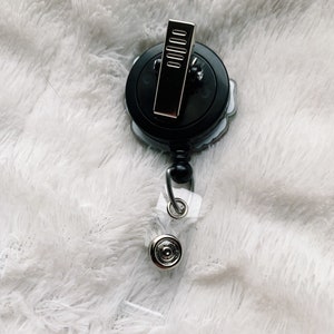 May include: A black and silver retractable badge holder with a floral design. The holder has a clip on the back and a clear plastic strap.
