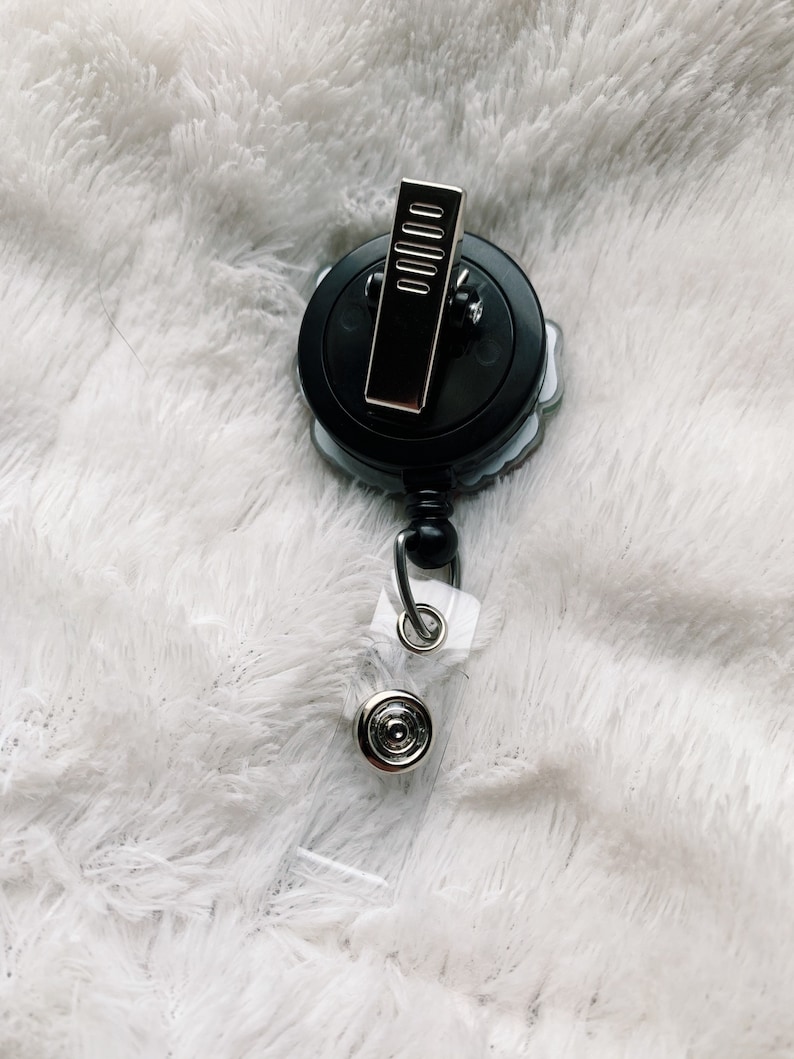 May include: A black and silver retractable badge holder with a floral design. The holder has a clip on the back and a clear plastic strap.