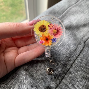 May include: A clear plastic badge holder with a round, floral design. The design features pressed flowers in shades of yellow, orange, pink, and purple.