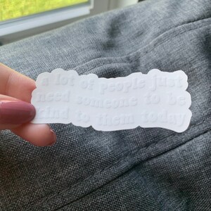 Op de afbeelding: Witte sticker met de tekst "veel mensen hebben gewoon iemand nodig die vandaag aardig tegen ze is".