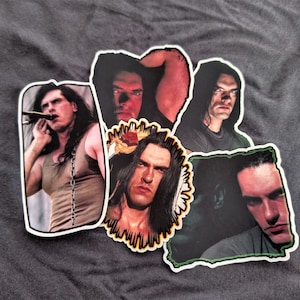May include: Five black and white stickers featuring portraits of men with long black hair. The stickers are on a gray surface.