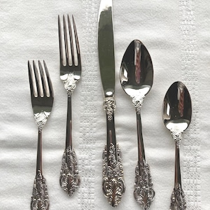 May include: A set of antique silverware, including two forks, a knife, and two spoons, arranged on a white tablecloth. The silverware has ornate, detailed handles and a shiny, reflective surface, suggesting a formal dining setting.