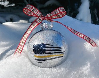 May include: A spherical silver glitter Christmas ornament featuring a black and white American flag graphic with a single horizontal gold stripe. The ornament is topped with a red, white, and black plaid fabric ribbon tied in a bow.