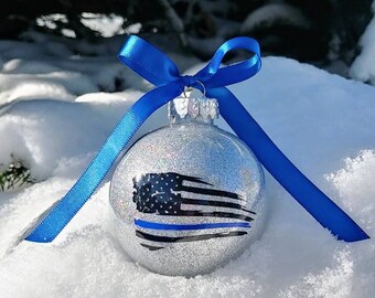 May include: A spherical silver glitter Christmas ornament featuring a black and blue American flag graphic. The ornament is topped with a metallic cap and a bright blue satin ribbon tied in a bow with two long tails hanging down the sides.
