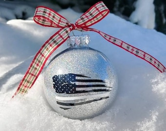 May include: A spherical silver glitter Christmas ornament featuring a black and white distressed American flag graphic with a single horizontal white stripe. The ornament is topped with a silver cap and a decorative red, green, and white plaid ribbon.