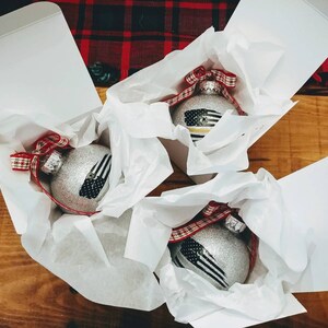 May include: Three silver glittery spherical ornaments, each nestled in a white gift box with white tissue paper. Each ornament features a black and white flag graphic with a thin colored stripe and is topped with a red and white plaid ribbon bow.