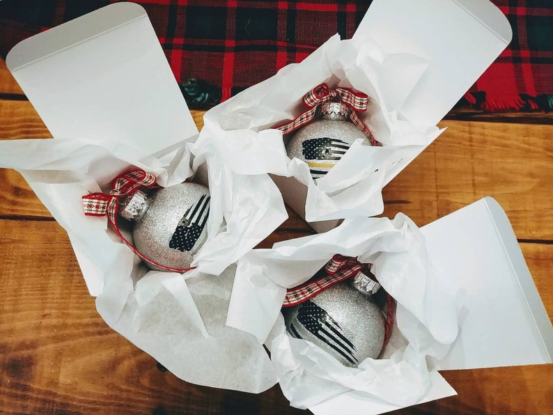 May include: Three silver glittery spherical ornaments, each nestled in a white gift box with white tissue paper. Each ornament features a black and white flag graphic with a thin colored stripe and is topped with a red and white plaid ribbon bow.