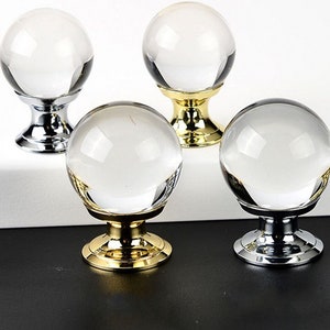 May include: Four spherical clear glass knobs with metallic bases. Two knobs feature polished silver-tone bases, while the other two feature polished gold-tone bases. Each knob has a round, transparent glass top attached to a circular pedestal mount.