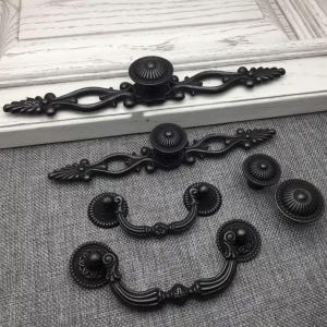 May include: A collection of matte black metal hardware including two long ornate drawer pulls with central knobs, two smaller swing-style bail pulls with circular backplates, and two round fluted knobs, all featuring intricate scrollwork details.