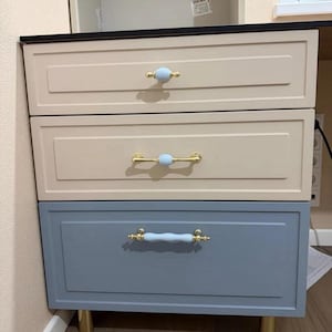 Pode incluir: Uma cómoda de três gavetas com duas gavetas superiores em acabamento bege claro e uma gaveta inferior em azul suave. Cada gaveta está equipada com um puxador de metal dourado acentuado por um centro de cerâmica azul claro ovalado.