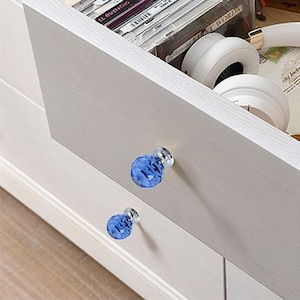 May include: A light-colored wooden drawer unit featuring two round, faceted blue glass knobs with silver-toned bases. The open drawer reveals a collection of compact discs and a pair of white over-ear headphones resting inside the storage space.