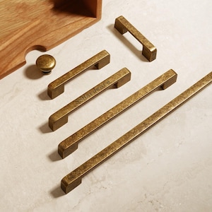 May include: A collection of antique brass cabinet hardware including four rectangular bar pulls of varying lengths and one round knob. One of the bar pulls features the engraved text Shardware. The hardware has a distressed, textured finish.