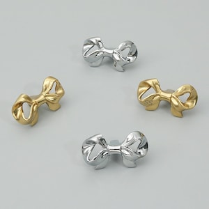 May include: Four metal cabinet knobs shaped like bows, arranged in a circular pattern. Two knobs have a polished silver finish, and two knobs have a gold finish. One gold knob features the letters SS engraved on the front surface of the bow.