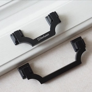 May include: Two matte black metal drawer pulls with a curved, geometric design and square mounting bases. The top pull features the white text SSHardware printed on the center bar. These hardware pieces have a solid, dark finish and angular edges.