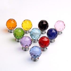 May include: A collection of ten spherical, faceted crystal cabinet knobs in various colors including orange, yellow, green, light blue, black, pink, purple, red, dark blue, and clear, each mounted on a circular silver-toned metal base.