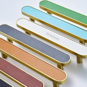 Puede incluir: Una colección de seis tiradores de armario alargados con marcos de metal dorado e incrustaciones texturizadas de colores en verde, azul claro, blanco, gris oscuro, bronceado y granate. El tirador blanco tiene el texto SSHardware.