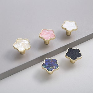 May include: Five flower-shaped cabinet knobs with gold-toned metal bases. The knobs feature decorative inserts in white, light pink, iridescent cream, dark blue with iridescent flecks, and solid black with subtle glittery specks.