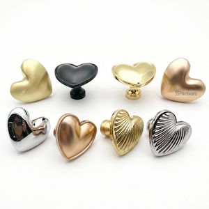 May include: A collection of eight heart-shaped cabinet knobs in various metallic finishes including matte gold, brushed black, polished gold, rose gold, chrome, and textured gold and silver, with the text SSHardware visible on one knob.