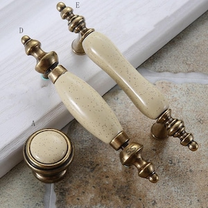 May include: Two elongated cabinet handles and one round cabinet knob, each featuring a cream-colored ceramic center with dark speckles and antique brass-toned metal ends. The handles are labeled with the letters D and E, and the knob is labeled A.