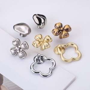 May include: A collection of seven metal cabinet knobs and pulls in various finishes including polished silver, brushed gold, and antique bronze. The hardware features clover-shaped designs, smooth rounded forms, and ring pulls with SSHardware text.