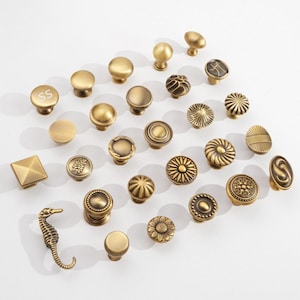 May include: A collection of brass-toned metal cabinet knobs and pulls in various shapes and textures. The assortment includes smooth round knobs, textured floral designs, geometric square shapes, and a seahorse-shaped pull, all featuring a metallic finish.