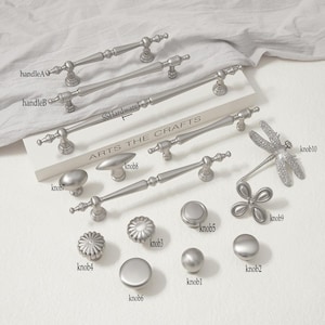 Puede incluir: Una colección de herrajes para armarios de metal plateado que incluye varios tiradores y pomos. Los artículos están etiquetados como handleA, handleB, knob1, knob2, knob3, knob4, knob5, knob6, knob7, knob8, knob9 y knob10. El texto SSHardware y ARTS THE CRAFTS es visible.