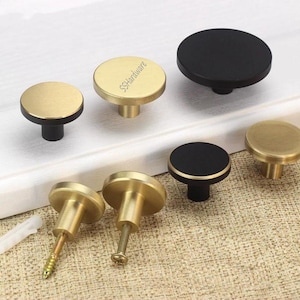 May include: A collection of round cabinet knobs in matte black and brushed gold finishes. Some knobs feature a flat circular top with a cylindrical base, while others show the threaded metal screw and plastic anchor attachment hardware.