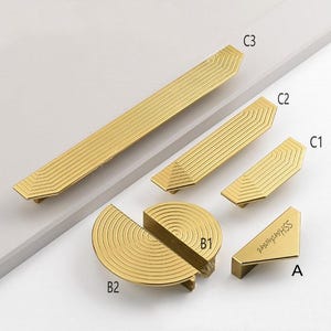 May include: A collection of gold-toned metal cabinet hardware including three linear pulls labeled C1, C2, and C3 with chevron-patterned ridges, two semi-circular knobs labeled B1 and B2 with concentric circular ridges, and a triangular knob labeled A.