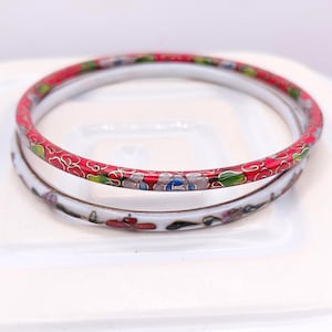 May include: Two circular bangle bracelets featuring cloisonné enamel work. The top bangle has a vibrant red background with multi-colored floral patterns in green, blue, and white. The bottom bangle is white with delicate red and green floral accents.
