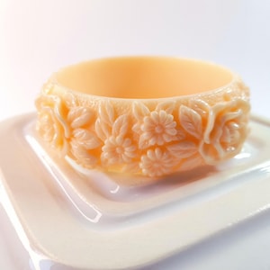 May include: A peach-coloured bangle bracelet with a floral design. The bracelet features carved roses, daisies, and leaves around the band. The bracelet is made of a solid material and sits on a white surface.