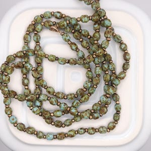 Puede incluir: Un collar largo con cuentas ovaladas de color verde oliva y azul claro, intercaladas con pequeñas cuentas de color bronce. El collar está colocado sobre una superficie blanca, con un cierre de color bronce visible.