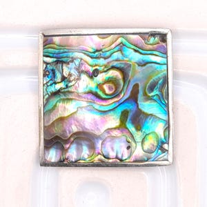 May include: A square-shaped brooch featuring a polished abalone shell inlay with iridescent swirls of blue, green, purple, and pink. The shell is encased in a smooth, silver-toned metal frame with clean, straight edges and a flat, uniform border.