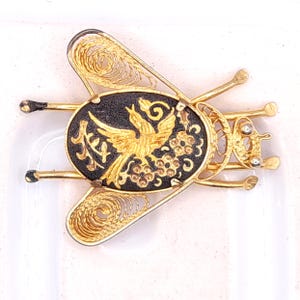 May include: A gold-toned insect-shaped brooch featuring filigree wings and a central oval black inlay. The inlay displays a gold-coloured bird and floral motif alongside Japanese kanji characters. The brooch has six thin legs and two antennae.