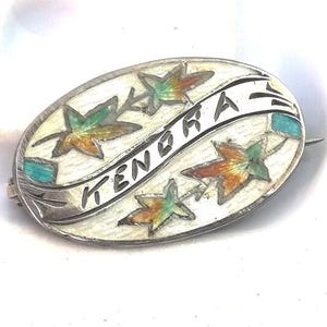 May include: An oval silver brooch with the name "KENORA" in black lettering. The brooch features colorful leaf designs in green, orange, and yellow, with small turquoise accents. The background is white.