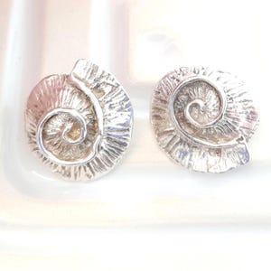 May include: A pair of silver-coloured earrings, each shaped like a spiral, reminiscent of an ammonite fossil. The earrings have a textured surface, with detailed ridges and curves. They are presented on a white surface, highlighting their metallic finish and design.