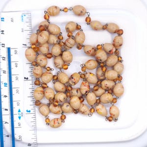 K&ouml;nnte beinhalten: Eine Halskette mit ovalen, beigen Perlen mit braunen und orangefarbenen Marmormustern, durchsetzt mit kleinen, durchscheinenden bernsteinfarbenen Perlen. Ein wei&szlig;es Lineal links zeigt eine L&auml;nge von etwa 38 cm an.