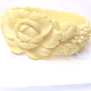 May include: Ivory-coloured bangle bracelet with a floral design. The bracelet features a large, carved rose with detailed petals and leaves. The edges of the bracelet have a scalloped design. The bracelet is made of a solid material.