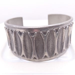 May include: A silver-toned metal cuff bracelet featuring a repeating embossed pattern of vertical, bone-shaped motifs. The background texture between each raised bone shape is finely stippled, creating a contrast against the smooth, polished metal.