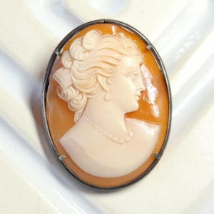 May include: Oval cameo brooch depicting a woman's profile, with intricate hair and a pearl necklace. The cameo is set on an orange background and framed by silver-coloured metal.