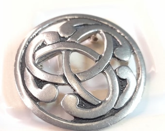 May include: A circular silver-toned metal brooch featuring an openwork design of an intertwined triquetra knot. The surface has a matte, brushed finish with darkened recessed areas that highlight the intricate, continuous looping pattern.