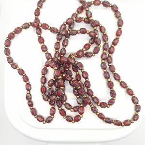 Puede incluir: Un collar largo con cuentas ovaladas de color rojo oscuro y espaciadores dorados. Las cuentas tienen un aspecto moteado y el collar está colocado sobre una superficie blanca. Es una joya.