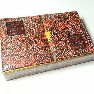 Könnte beinhalten: Eine dekorative Box mit einem rot, schwarz und weiß gesprenkelten Muster. Die Box hat goldfarbene Verzierungen und den Text "WITH THE COMPLIMENTS OF HOLLIS BROS LTD". Ein goldener Quaste ist sichtbar. Die Box ist teilweise geöffnet.