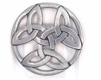 May include: A circular silver-toned metal brooch featuring an intricate, openwork triquetra knot design. The piece has a smooth, polished finish with recessed areas that highlight the interlacing pattern of the three-pointed Celtic knot motif.