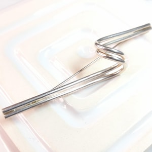 May include: A silver brooch with an abstract design. The brooch has a pin and a series of curved and straight metal elements, creating a modern, sculptural appearance. The brooch is set against a white background.