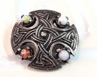 Puede incluir: Broche de cruz celta plateada con intrincado diseño de nudos. El broche presenta cuatro gemas redondas y coloridas: verde, naranja y dos tonos de morado. Un accesorio decorativo.