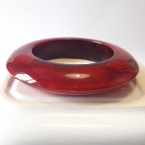 Puede incluir: Un brazalete ovalado brillante en un rico tono marrón rojizo. El brazalete tiene una superficie lisa y pulida y un interior oscuro. El diseño es simple y elegante, adecuado para el uso diario o ocasiones especiales.